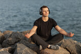 Healthy sport man meditates listening to relaxing music while sitting on stones at coast