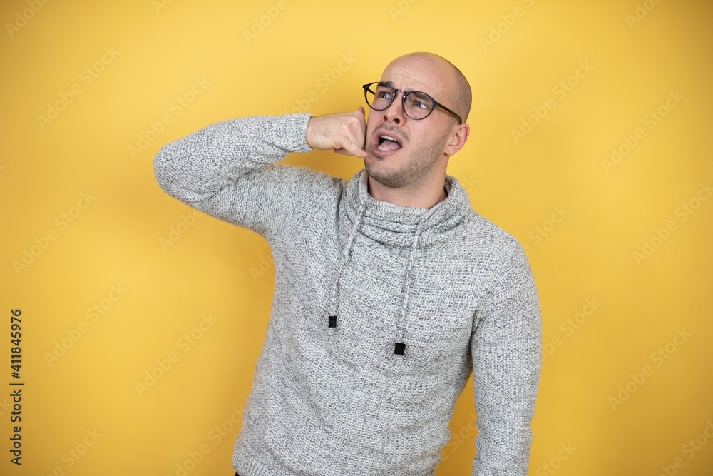 Young bald man wearing glasses over yellow background confused doing phone gesture with hand and fingers like talking on the telephone Young bald man wearing glasses over yellow background confused doing phone gesture with hand and fingers like talking on the telephone