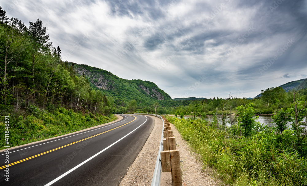 Canadian road in Quebec Province meandering
 between river and and cliffs with trees everywhere at summer time during a cloudy day