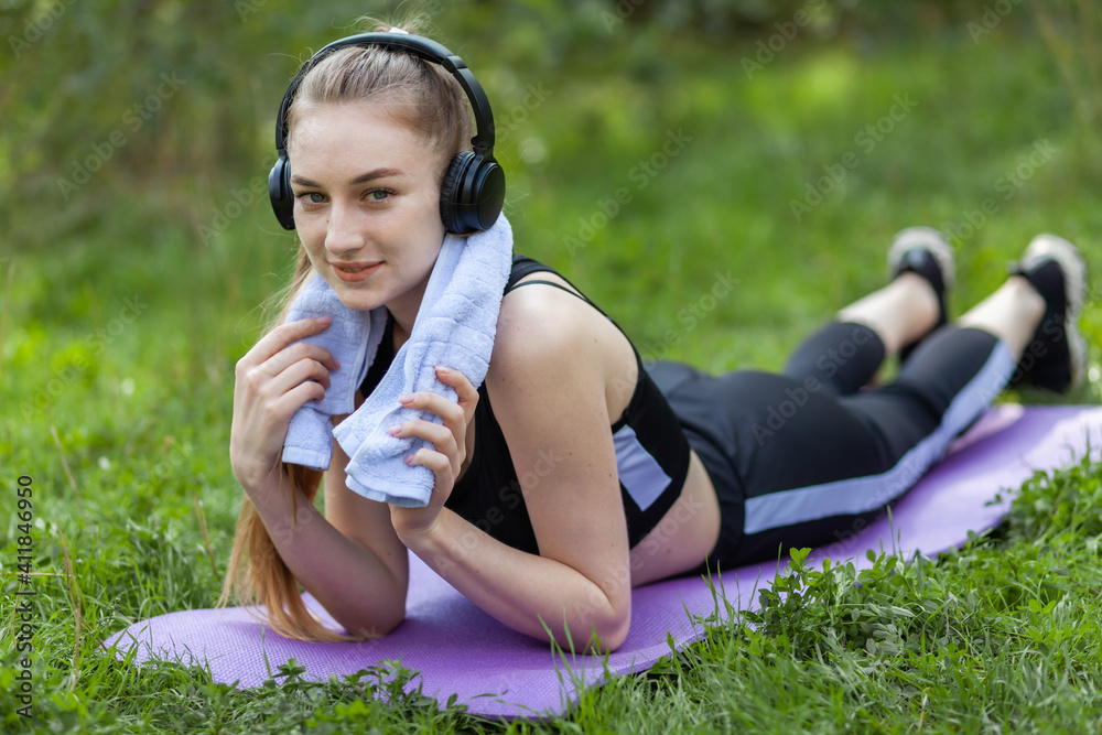 Fit slim woman with a towel on her shoulders lies relax on a mat and listen music in headphones at park. Healthy lifestyle Fit slim woman with a towel on her shoulders lies relax on a mat and listen music in headphones at park. Healthy lifestyle