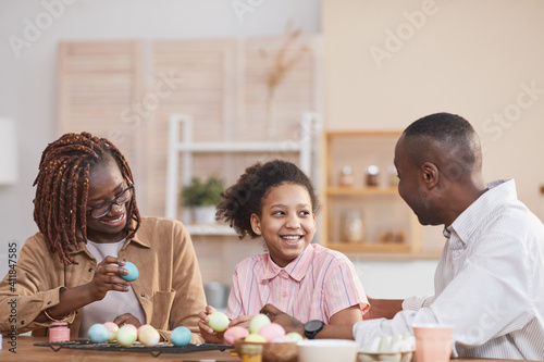 Portrait of laughing African-American family painting Easter eggs together while sitting at wooden table in cozy home interior and enjoying DIY art