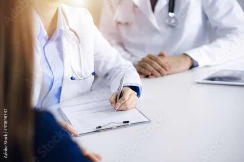 Unknown woman-doctor with male colleague are consulting patient woman while sitting at the desk in sunny clinic, close-up. Covid 2019