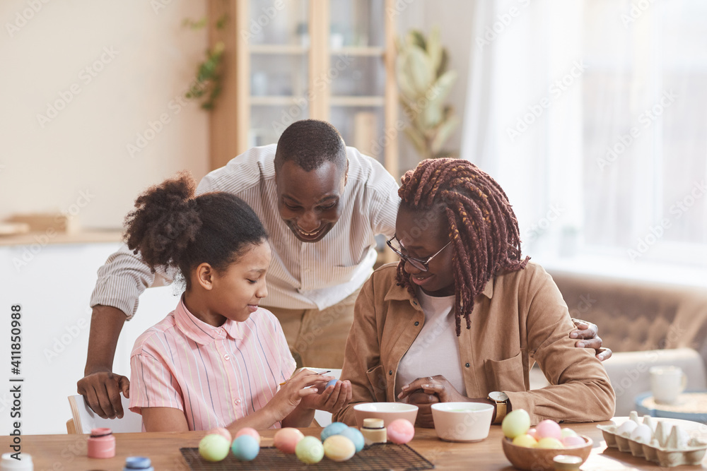 Portrait of loving African-American family painting Easter eggs together while sitting at wooden table in cozy home interior and enjoying DIY art, copy space Portrait of loving African-American family painting Easter eggs together while sitting at wooden table in cozy home interior and enjoying DIY art, copy space