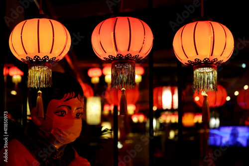 Red Chinese paper lanterns glow in a row on a night street on restaurant entrance and statue in medical mask background. New Year celebration in China during covid-19 coronavirus pandemic