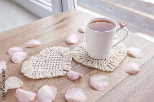 Macrame handmade Hobby. Tea in a cup on white macrame coaster on wooden table with rose petals. Food stylist. Eco macrame home decoration. St. Valentine's Day