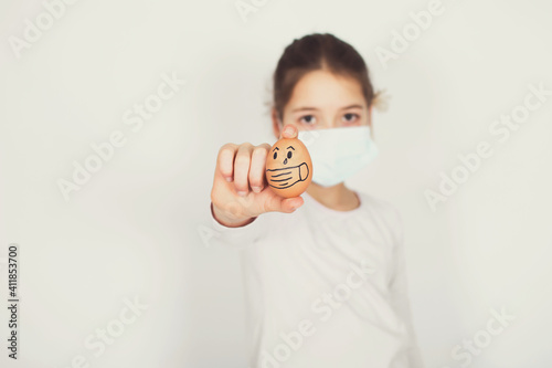 Young girl with face mask is holding painted egg with face mask isolated on white background. Easter, coronavirus and social distance concept.
