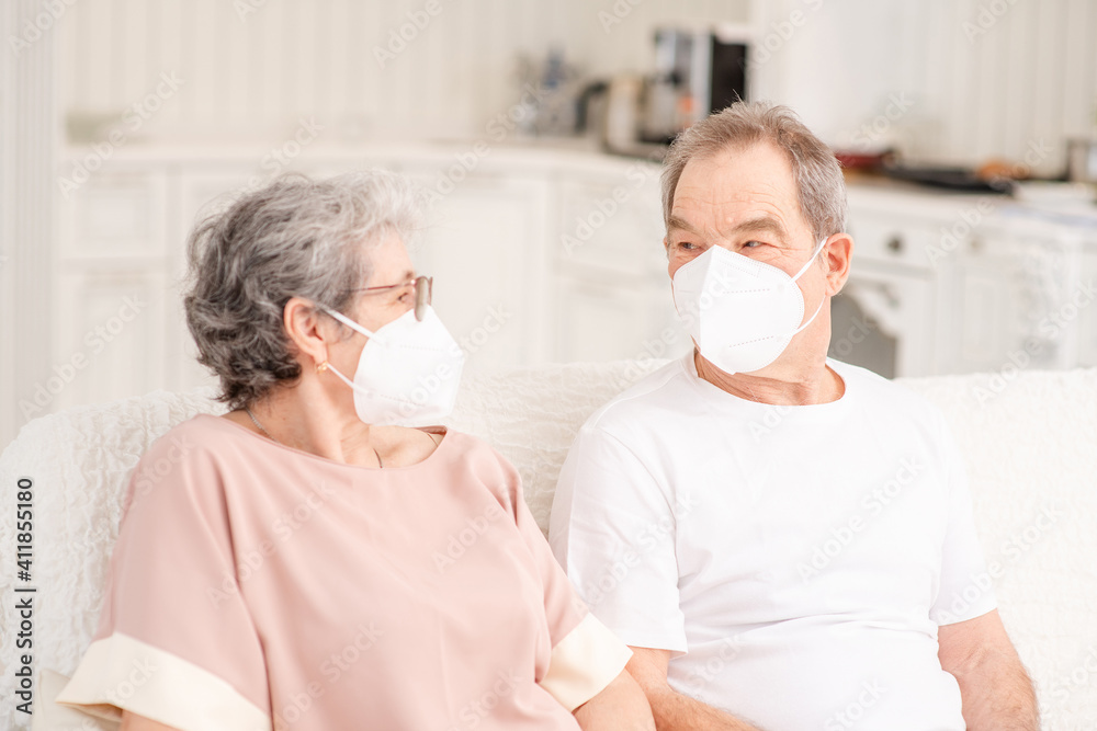 An elderly couple with medical masks sit on the couch at home and chat during a pandemic. Ways not to get bored during lockdown and develop while sitting at home.