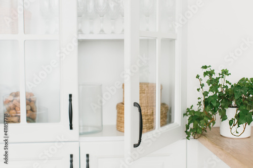 Opened door of white glass cabinet with clean dishes and decor. Scandinavian style kitchen interior. Organization of storage in kitchen. Selective focus.