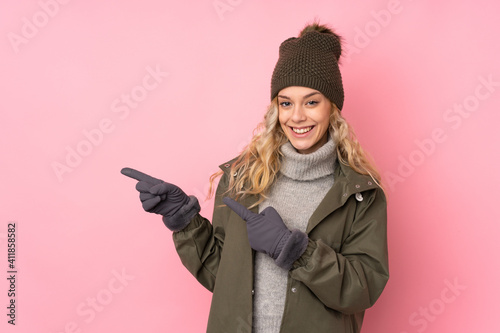 Young girl with winter hat isolated on pink background pointing finger to the side