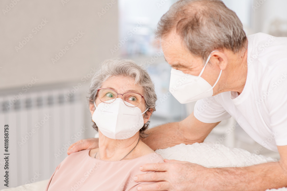 An elderly couple with medical masks sit on the couch at home and chat during a pandemic.
