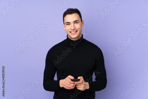 Young caucasian handsome man with turtleneck sweater isolated on purple background sending a message with the mobile