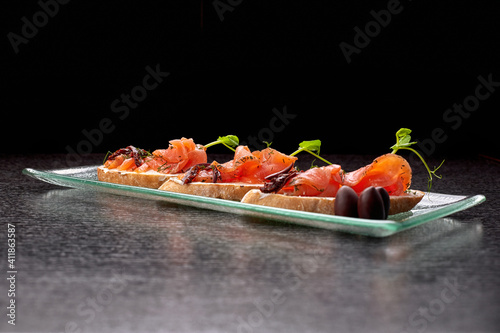Bruschetta with trout, salmon, cream cheese and microgreen on a glass plate on a black background
