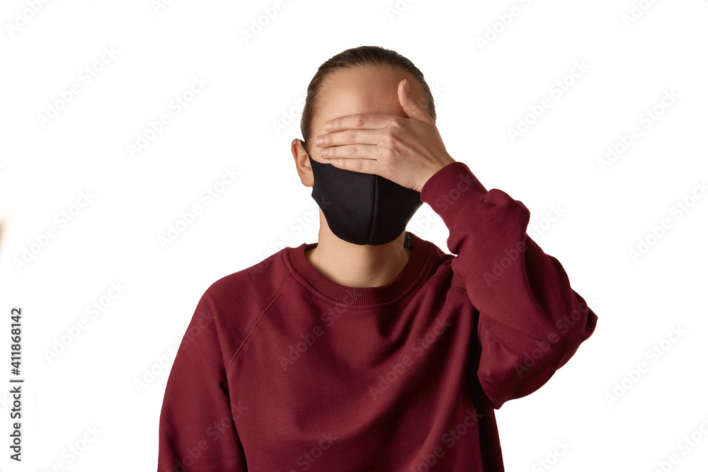 Tired young european woman wears black face mask and burgundy sweatshirt, closes eyes with her hand in disbelief, doesn't want to see something, isolated over white background. Tired young european woman wears black face mask and burgundy sweatshirt, closes eyes with her hand in disbelief, doesn't want to see something, isolated over white background.