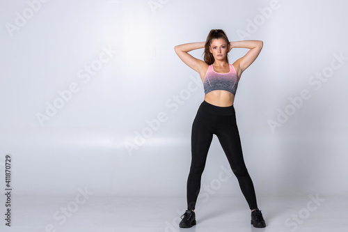Athletic fitness woman in sportswear standing crossing her arms behind her head on white background. Trainer stretching her core muscles in a standing pose  