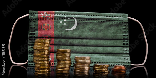 Medical mask with the flag of Turkmenistan behind some shrinking stacks of various coins.(series) photo