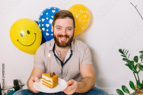 Bearded man with honey cake celebrates his birthday alone sitting on the floor in the house. Balloons. Sports Equipment. Lifestyle. Money tree in a pot. Isolation concept photo