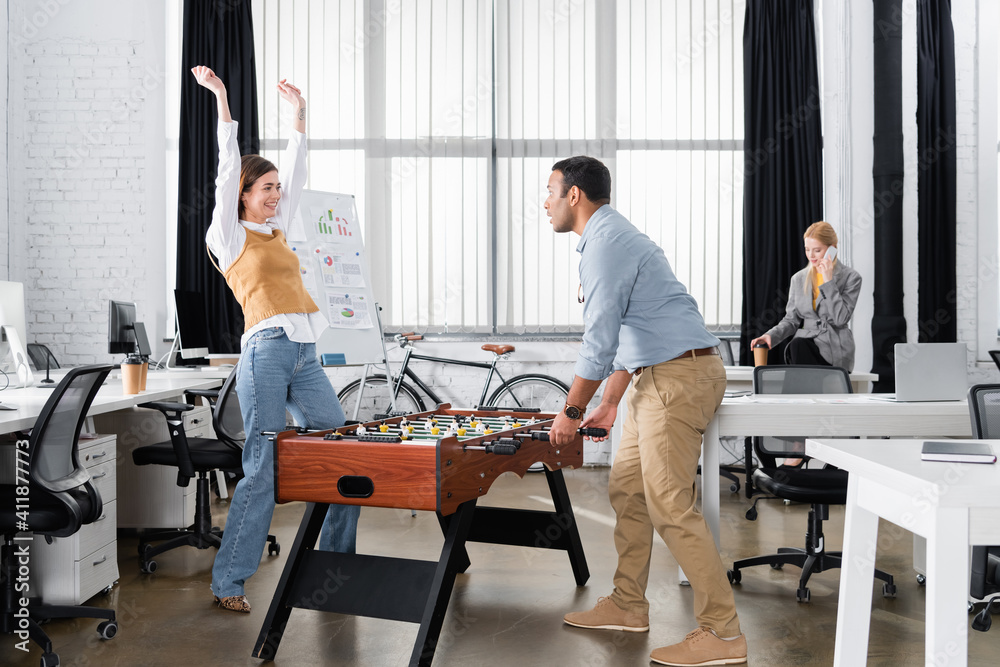 Happy businesswoman looking at indian colleague near table soccer in office