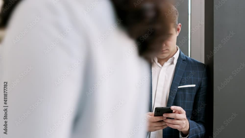 Panning of young handsome businessman wearing checkered suit standing in business center hall, using his cellphone then looking at camera and smiling Panning of young handsome businessman wearing checkered suit standing in business center hall, using his cellphone then looking at camera and smiling