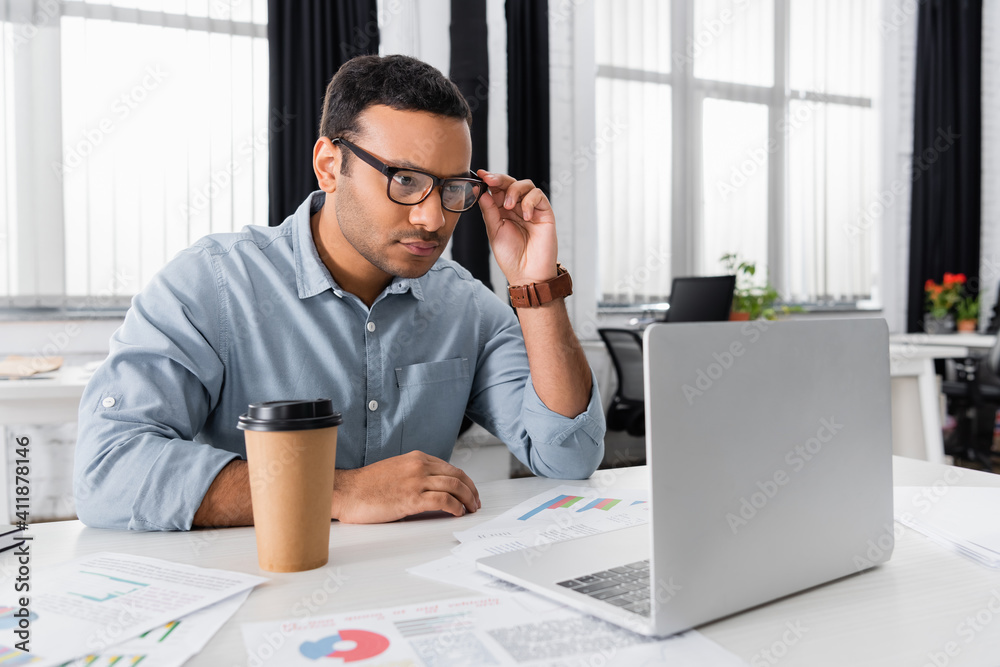 Obraz premium Indian businessman looking at laptop near coffee to go and papers on blurred foreground