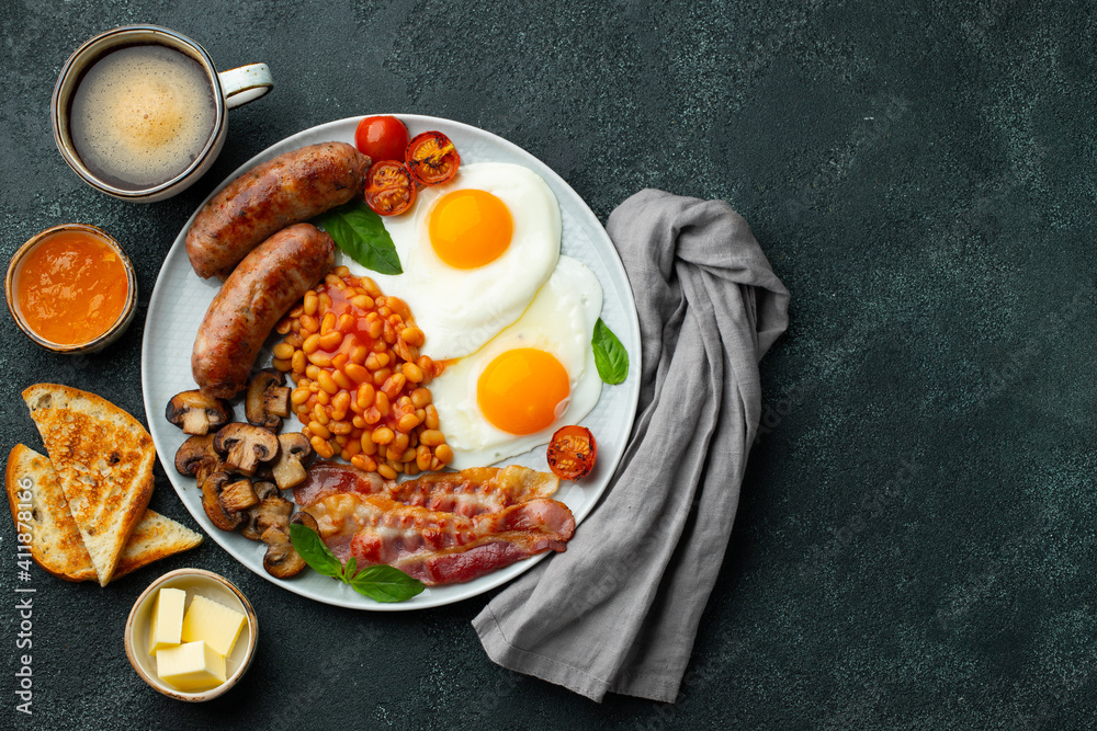 Full English breakfast on a plate with fried eggs, sausages, bacon, beans, toasts and coffee on dark stone background. With copy space. Top view Full English breakfast on a plate with fried eggs, sausages, bacon, beans, toasts and coffee on dark stone background. With copy space. Top view