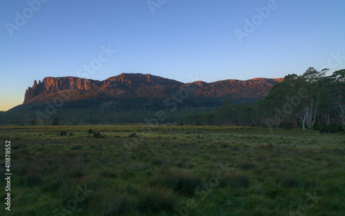 Wide green grassland, Eucalyptus trees and illuminated steep and rocky mountain ridge at sunset, The Overland Track, Tasmania, Australia