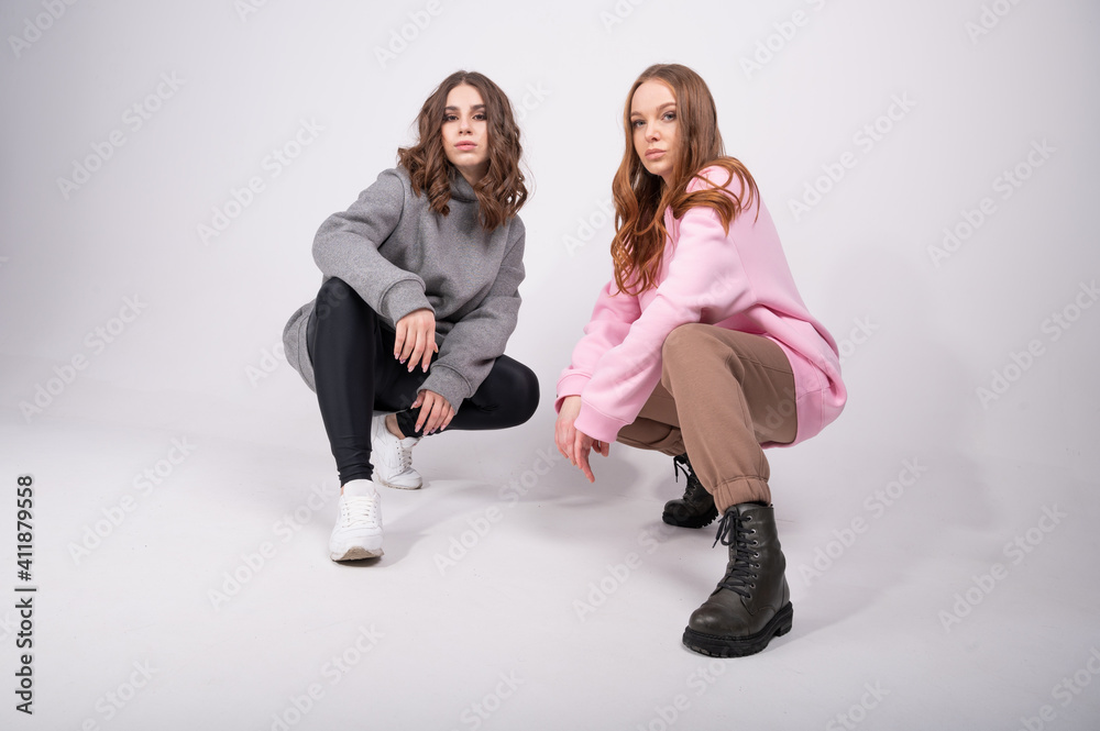 Naklejka premium Full length view of two young girls in sports fashionable clothes sit on their haunches on a white background. People fashion lifestyle concept.