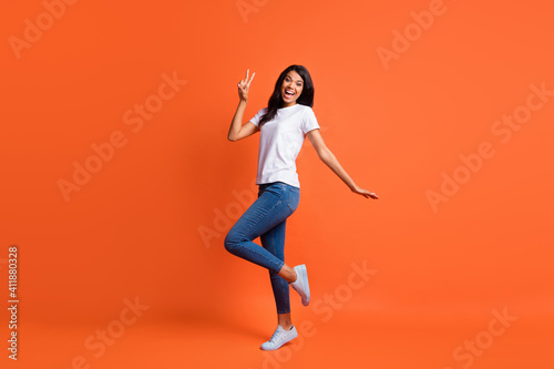 Full size photo of optimistic girl stand show v-sign wear white t-shirt jeans sneakers isolated on orange color background