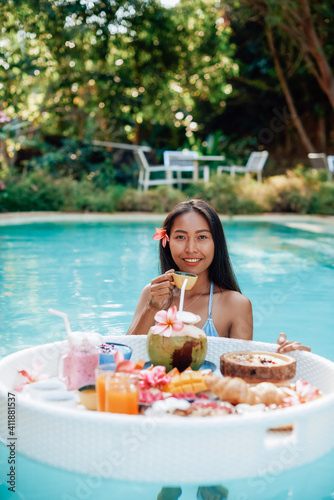Asian woman and asian food. Young woman with dark hairs poses in swimming pool with floating table and cup of juice. © Fxquadro