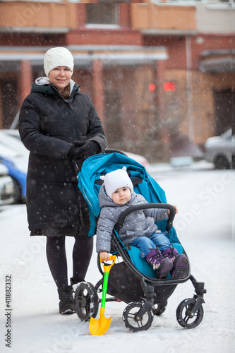 Young mother standing with her toddler child sitting in baby carriage, full-length portrait outdoor at winter time