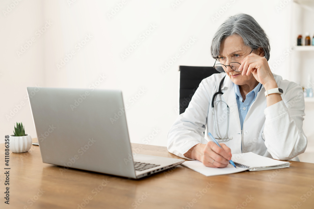Senior old female doctor with a stethoscope in a lab coat and glasses sitting at the desk and looking at the laptop, studying online, freshening-up her memory, and making notes in the notebook Senior old female doctor with a stethoscope in a lab coat and glasses sitting at the desk and looking at the laptop, studying online, freshening-up her memory, and making notes in the notebook