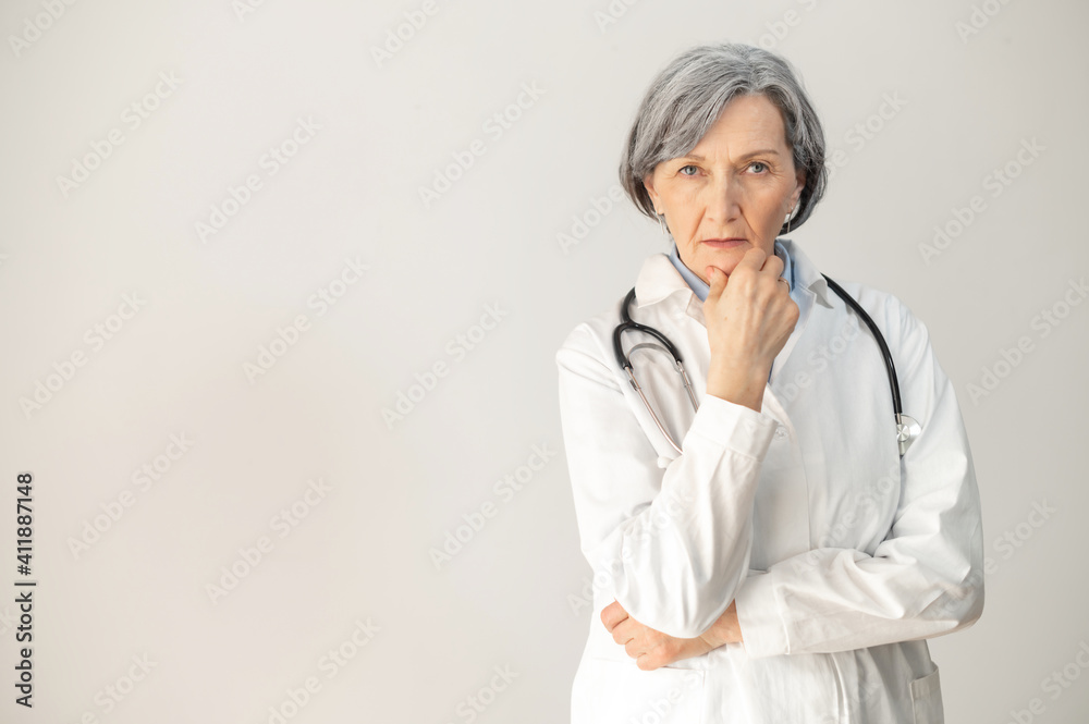 Portrait of a senior mature female doctor standing with a stethoscope and posing, isolated over gray background, wearing a medical robe, placed her hand on a chin and confidently looking at the camera Portrait of a senior mature female doctor standing with a stethoscope and posing, isolated over gray background, wearing a medical robe, placed her hand on a chin and confidently looking at the camera