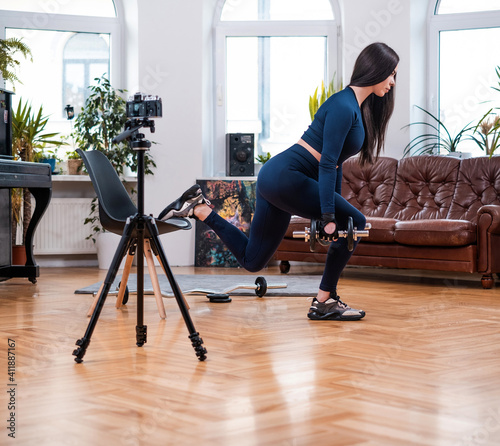 Beautiful and stylish sportswoman in blue clothing exercises with chair and dumbell posing on camera in modern apartment with furniture.