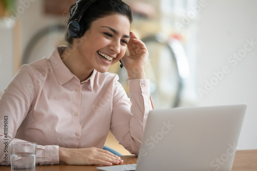 Happy consultant in headphones using laptop for video call and laughing. Female tutor teaching class online from home, holding webinar, smiling at webcam. Young Indian operator giving client support