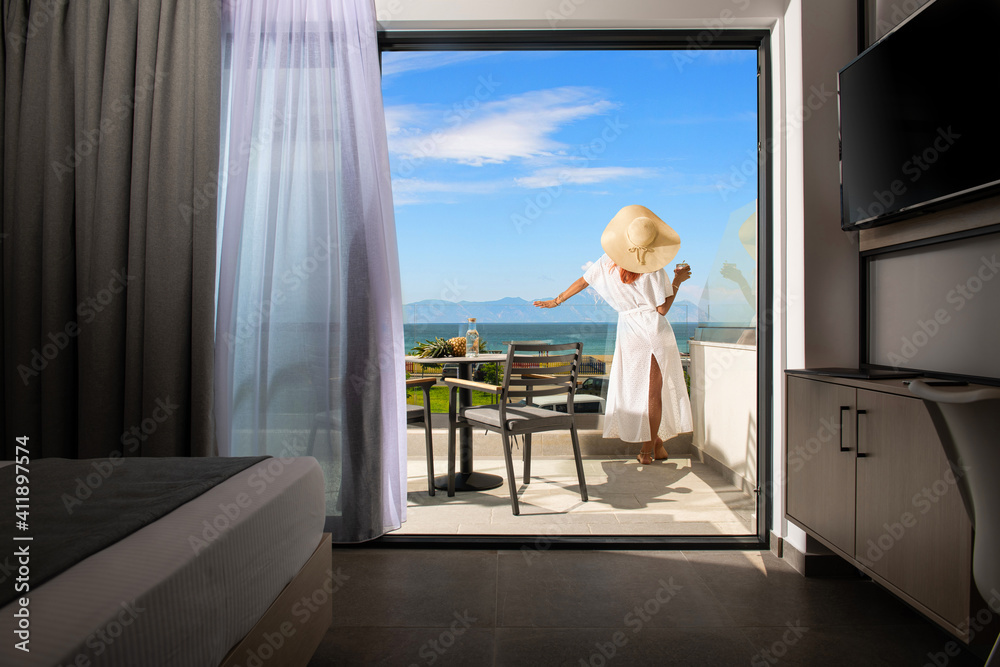 Back view of young woman in white summer dress and straw hat on open balcony terrace of resort hotel room with sea view landscape