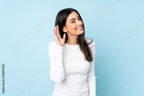 Young caucasian woman isolated on blue background listening to something by putting hand on the ear photo