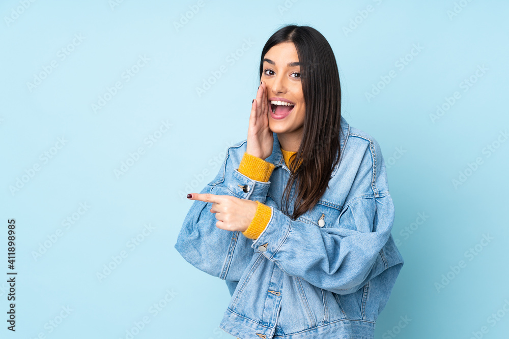 Young caucasian woman isolated on blue background pointing to the side to present a product and whispering something Young caucasian woman isolated on blue background pointing to the side to present a product and whispering something