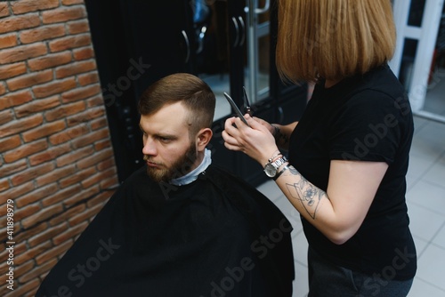 Stylish man sitting barber shop Hairstylist Hairdresser Woman cutting his hair Portrait handsome happy young bearded caucasian guy getting trendy haircut Attractive barber girl working serving client