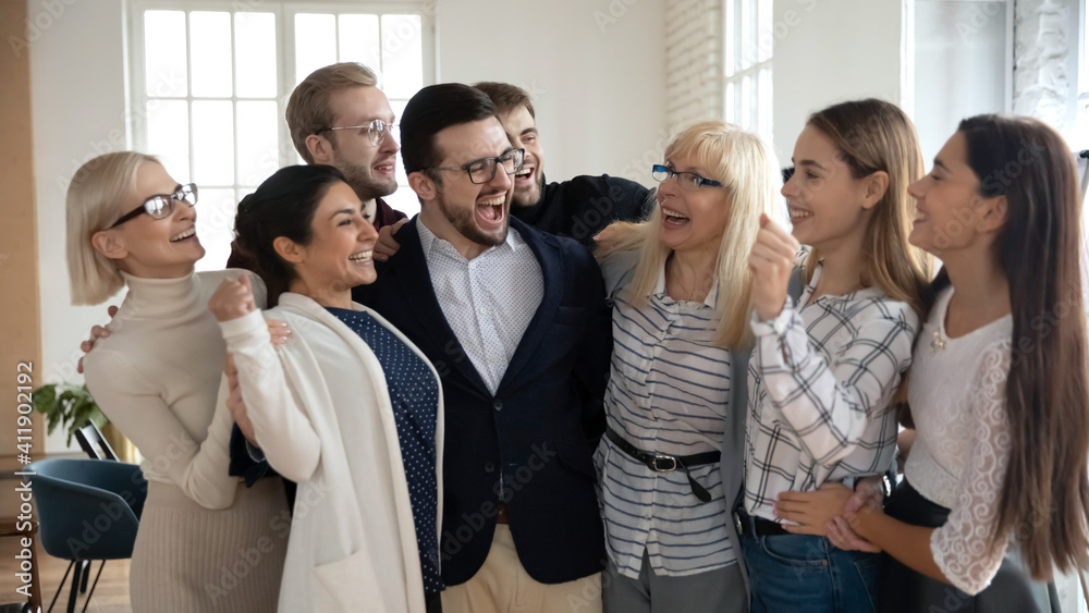 Group of happy workers of different generations hugging like family together, laughing. Excited diverse business team cheer celebrating success. Corporate achievement, win, teamwork, community concept