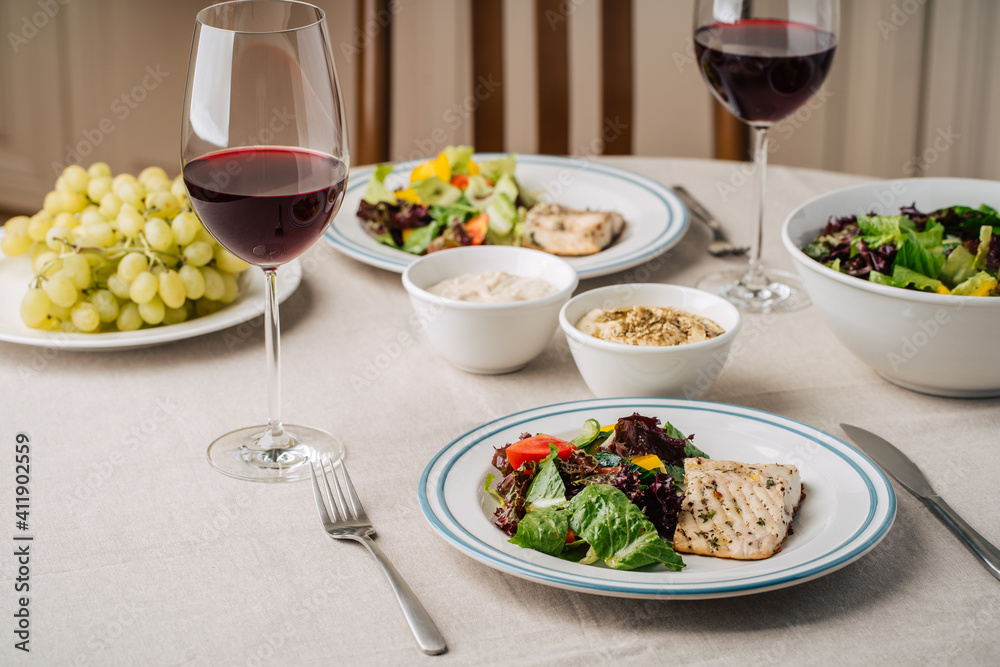 White fish steak with vegetable salad, sauce, hummus, grapes and glasses of wine. Served for two in the kitchen White fish steak with vegetable salad, sauce, hummus, grapes and glasses of wine. Served for two in the kitchen
