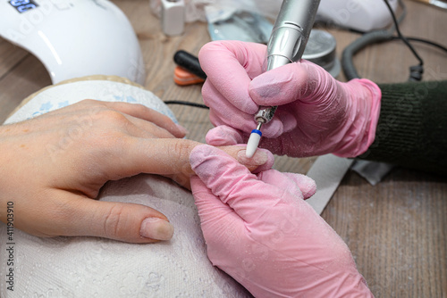 Removing the old hybrid nail using an electric nail grinder, the beautician has pink latex gloves.