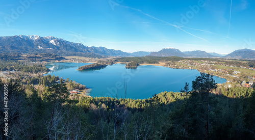 A drone shot of a Faaker lake in Austrian Alps. The lake is surrounded by high mountains. There is a small island in the middle. Green forest growing at the shore. Clear and sunny day. Calmness © Chris