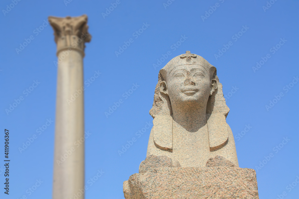 A sphinx in the northern Egyptian city of Alexandria with a column in the background. An ancient figure in the form of an animal with a human head. It is a clear day with blue skies.