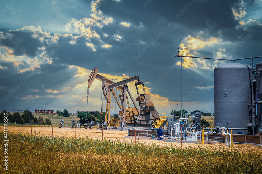 Two pump jacks and tanks and oil field equipment in fenced area in pasture under dramatic skies with houses on hill on horizon - summer Two pump jacks and tanks and oil field equipment in fenced area in pasture under dramatic skies with houses on hill on horizon - summer