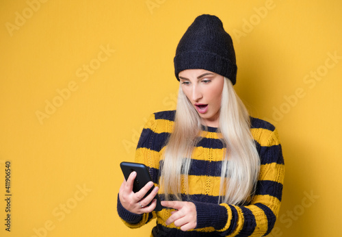 Astonished girl with smartphone, model wearing woolen cap and sweater, isolated on yellow background. Trendy woman. Unbelievable message