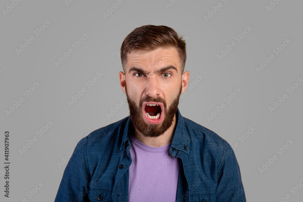 Portrait of young bearded guy shouting furiously, expressing his anger over grey studio background. Negative emotions Portrait of young bearded guy shouting furiously, expressing his anger over grey studio background. Negative emotions
