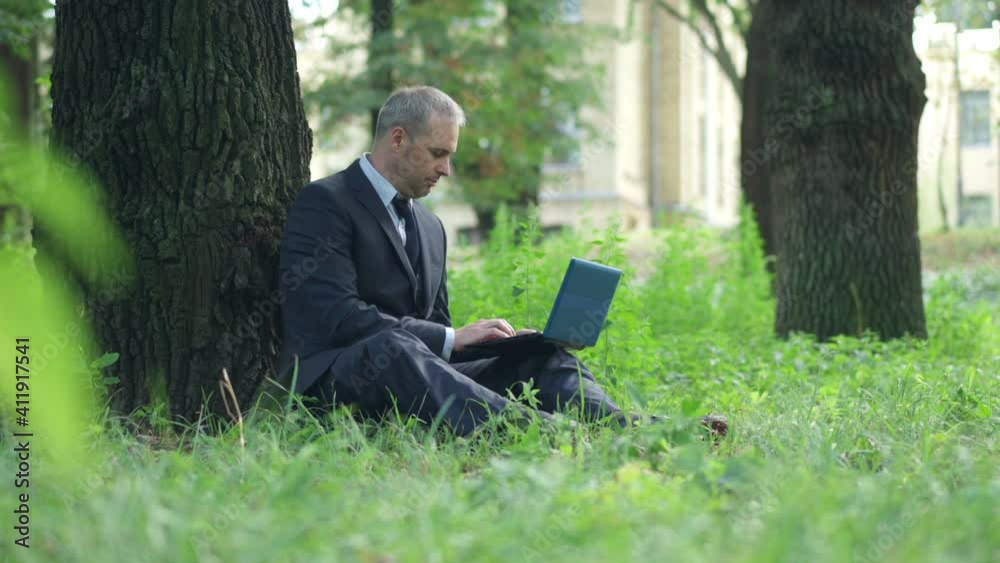 Wide shot of relaxed Caucasian businessman typing on laptop sitting on green summer meadow on sunny day. Side view of confident focused middle aged man working online outdoors. Freelance and business. Wide shot of relaxed Caucasian businessman typing on laptop sitting on green summer meadow on sunny day. Side view of confident focused middle aged man working online outdoors. Freelance and business.