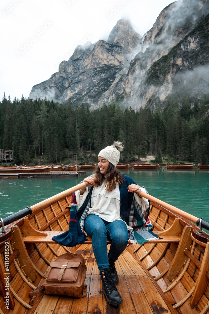 Happy tourist woman with hat sitting in a wooden boat on Braies lake surrounded by the mountains of the Italian Alps 