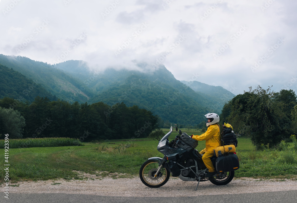 A girl in a yellow raincoat, shoe covers and helmet. Motorcyclism and travel. Sightseeing tour. Top of the Mountains. A gray day with clouds. Copy space, biker's outfit. High resolution panorama