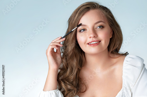 Young girl using brush for eyebrows. Photo of woman with perfect makeup on light blue background. Beauty and Skin care concept. Plus size model.
