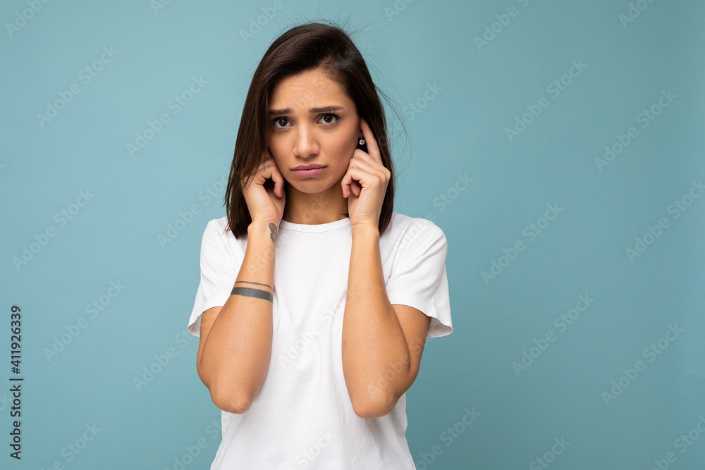 Portrait of sad sorrowful young pretty nice brunette woman with sincere emotions wearing casual white t-shirt for mockup isolated on blue background with copy space and covering ears with fingers Portrait of sad sorrowful young pretty nice brunette woman with sincere emotions wearing casual white t-shirt for mockup isolated on blue background with copy space and covering ears with fingers